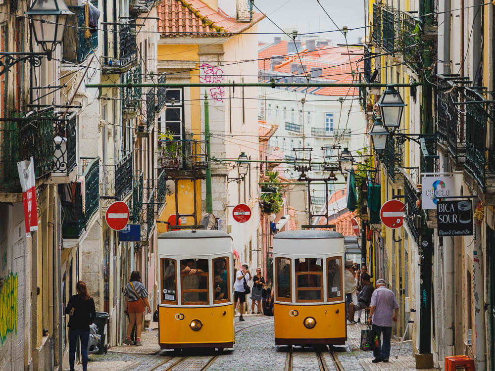 Two white and yellow trams going up a hill in Lisbon. There are wires connecting them to the electricity, some people walking near it, and some signs. On both sides of the street, there are houses with balconies.