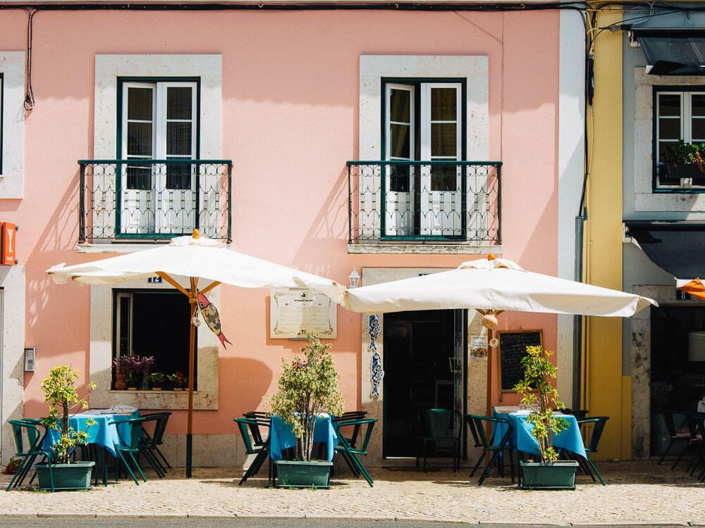 A pink building with two small balconies and three tables set in front of it, with umbrellas and a small tree in front of each table.  