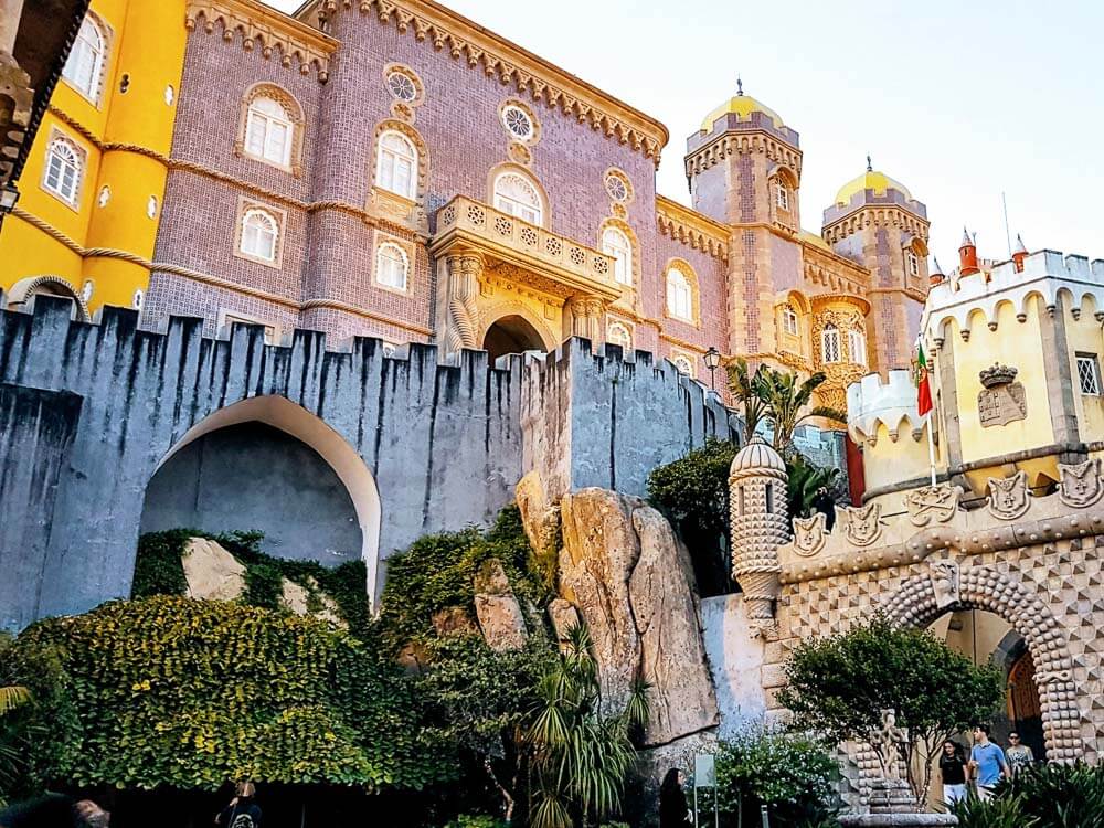 The colorful Pena Palace, with a grey wall above some greenery, and an ornate archway to the right. Above, there’s a pink construction and, to the left, a yellow piece of wall.