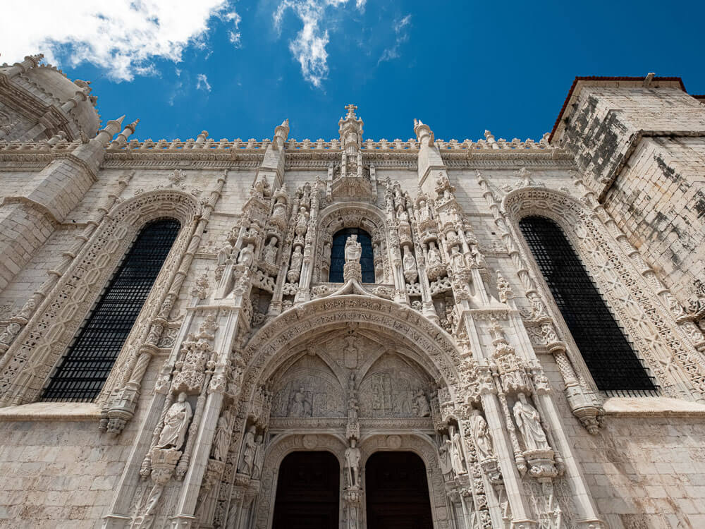 View from bellow of the entrance of the Jerónimos Monastery. The construction is white and there’s a big arch with ornate decorations and two tall windows on each side. 