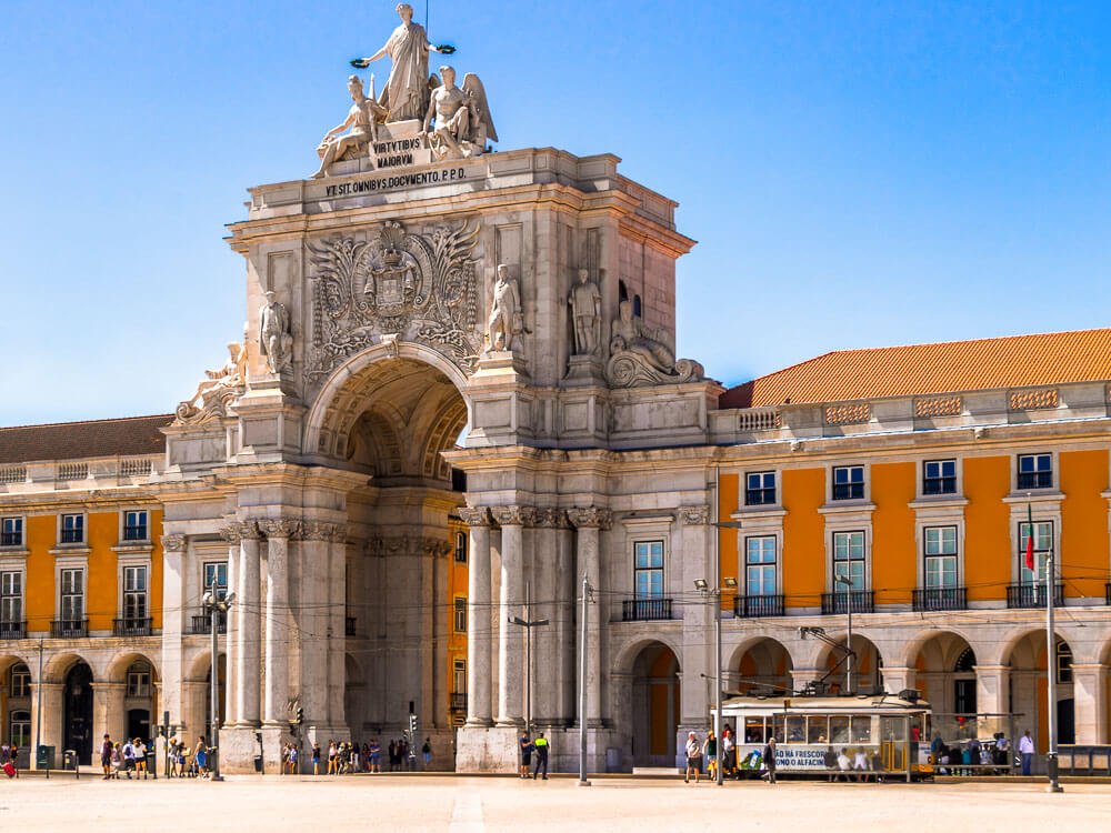 Commerce Square in Lisbon, with the Augusta Arch in the middle and yellow buildings with smaller arches beneath them on each side. There are people near the arch and a tram.