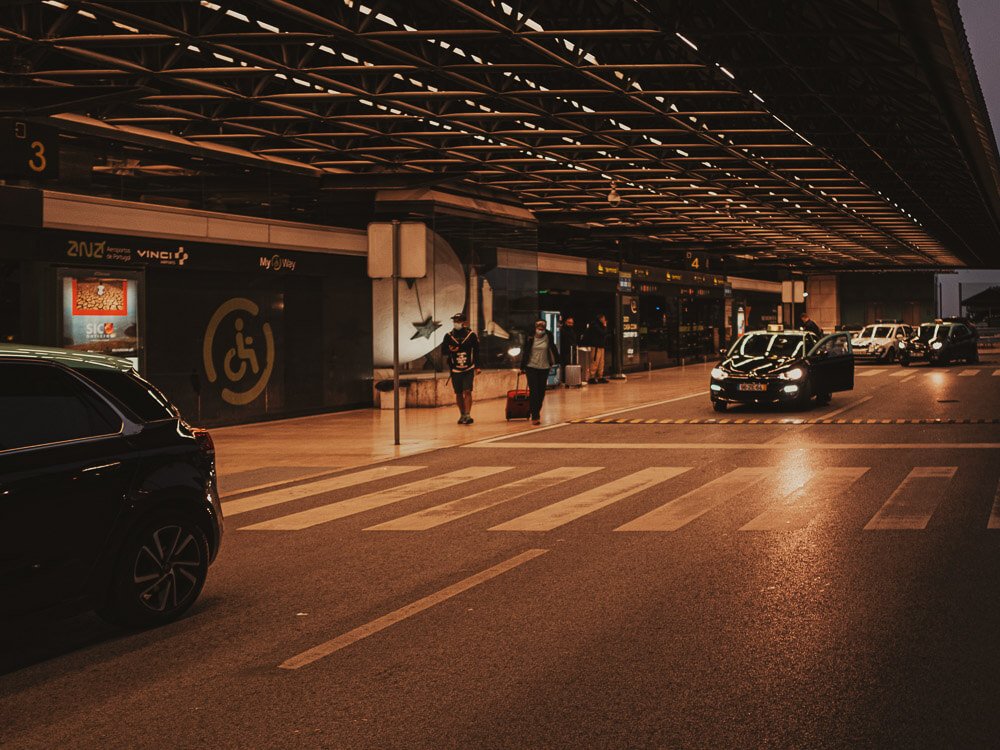 Lisbon Airport at night. There are taxis driving down the road, a metallic structure as the ceiling, two people walking and a crosswalk.