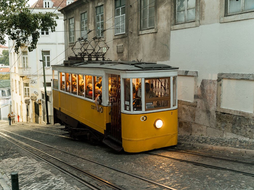 A yellow and white tram going up a hill in Lisbon. It’s full of people. The street is cobblestone and it’s possible to see the tracks.