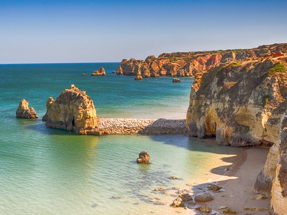 A beach in the Algarve, Portugal, with clear turquoise waters and surrounded by cliffs, which are casting some shadows on the sand. There are a couple of sea stacks on the water.