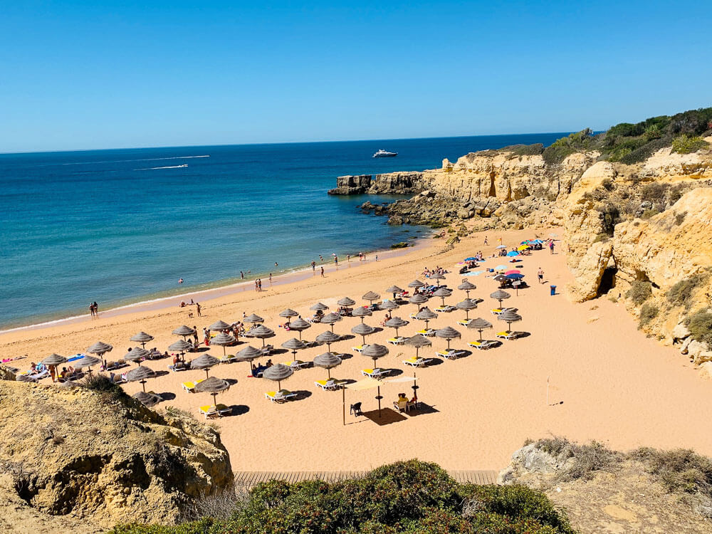 A beach in Portugal surrounded by clifs, with calm waters and numerous straw sun umbrellas and yellow sunbeds set up on the sand, forming 4 lines. There are a couple of colorful umbrellas to the right, and some people under the shade and on the water.