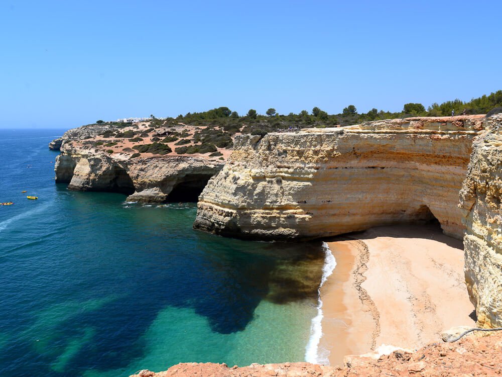 A beach in the Algarve, Portugal, surrounded by high cliffs, which have some vegetation on top. The ocean is turquoise and there are a couple of caves beneath the cliffs. 