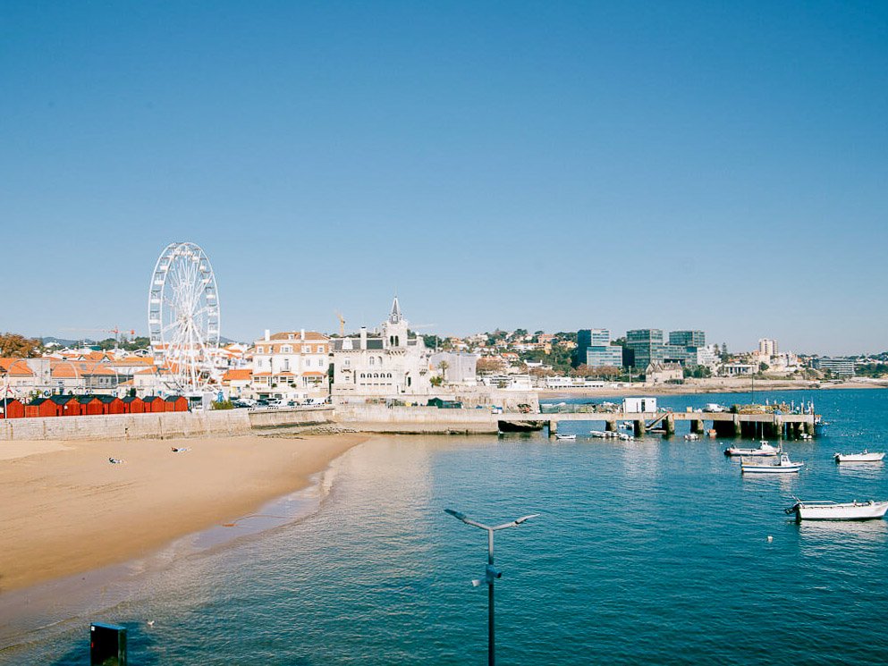 A beach in Cascais, Portugal, with a pier and a ferris wheel. The shoreline is surrounded by buildings and there are a couple of boats on the water.