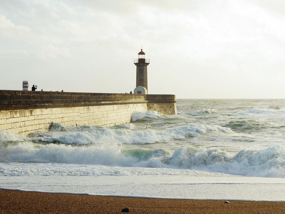 A beach in Northern Portugal. The sand is thick, and the ocean is rough, with some waves breaking. There’s a pier on the left with a lighthouse. The sky is cloudy.