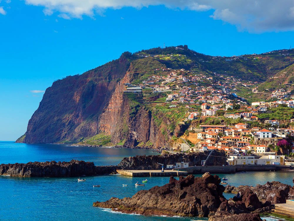 Madeira Island, where there’s a cliff with houses built on it and lots of underbrush. There’s a pier going into the sea and some rocks on the water. The ocean is calm and the sky is clear.