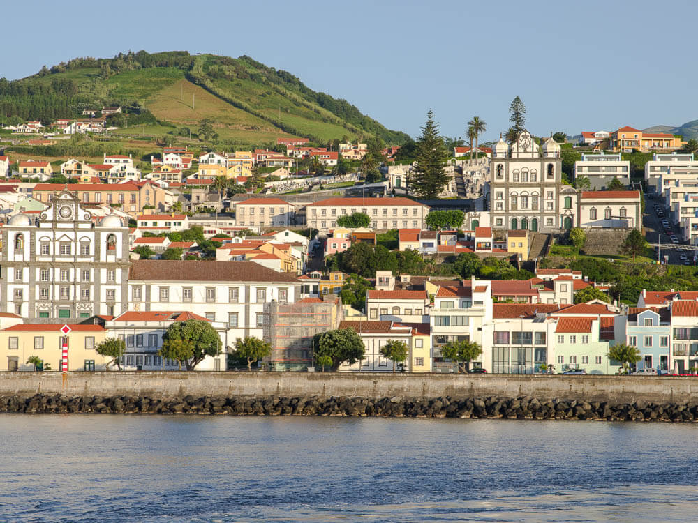 A town in the Azores, with houses built along the shoreline, various trees and a small hill on the background. The ocean in front of it is calm. There’s a wall lined with rocks separating the houses from the beach.