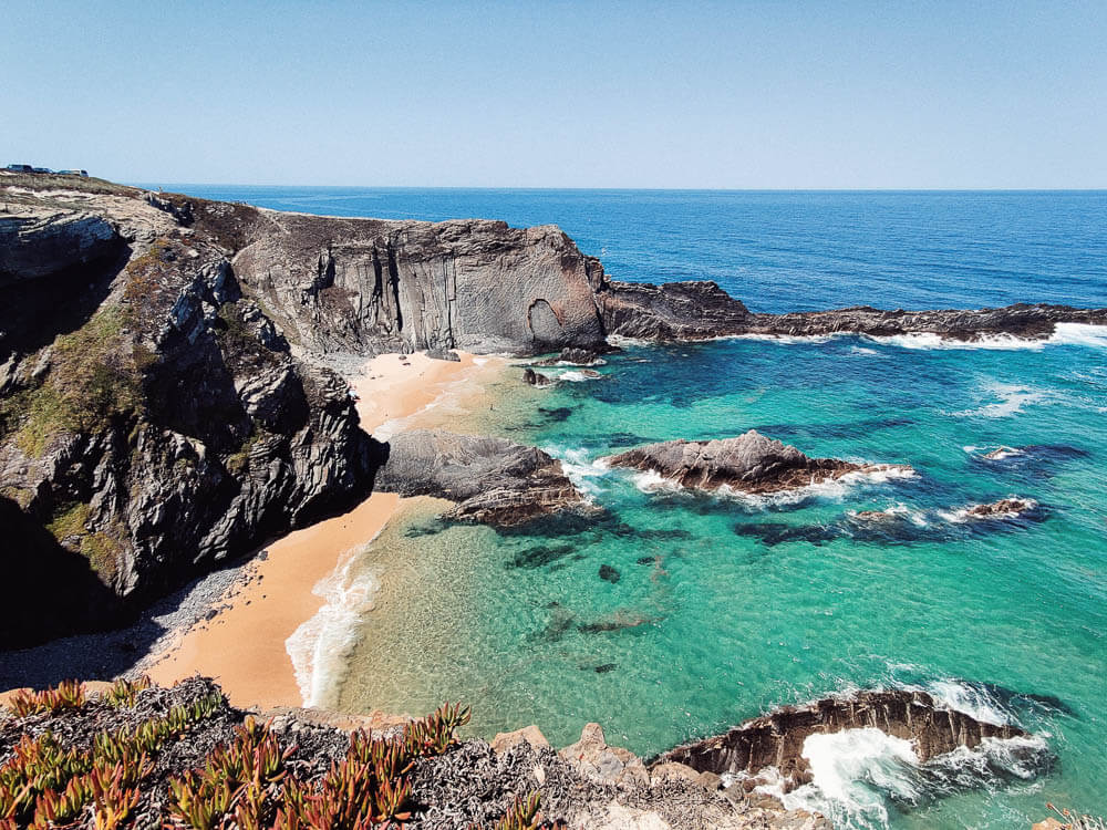 A beach in the Alentejo shoreline, with rugged rocky grey cliffs and underbrush. There are rocks inside the water. The ocean is very clear and turquoise.