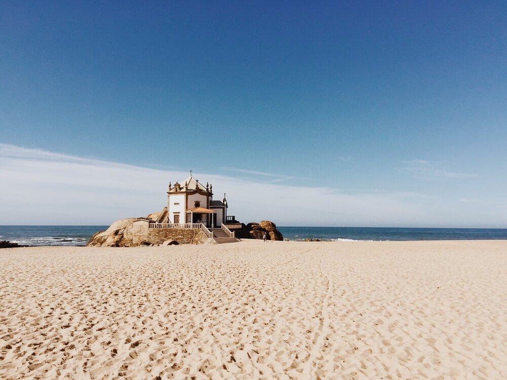 A long stretch of white sand beach in Northern Portugal, with the ocean in the background. In the middle, more to the left, there’s a chapel with a white picket fence, surrounded by rocks. The sky is clear, with some clouds to the left. 