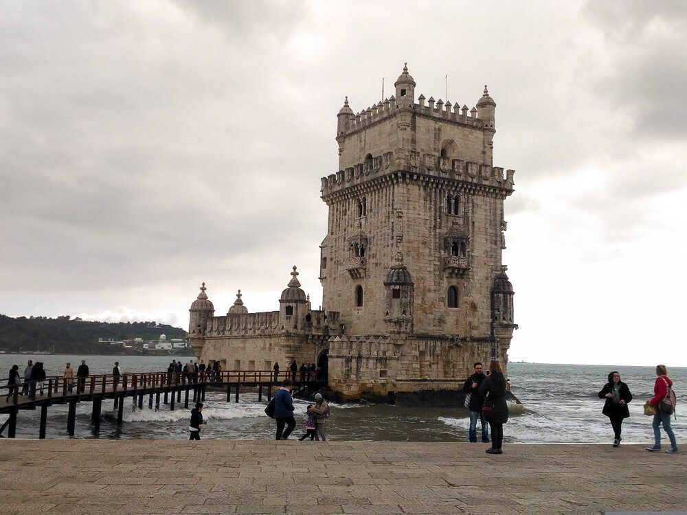 People walking around the Belém Tower, rising from the Tagus River. The sky is cloudy and people are wearing winter clothes.