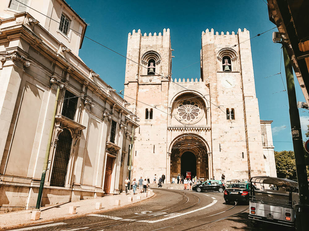 People walking near Lisbon Cathedral, which has two towers with bells and a round stained glass window above an arched door. There are three cars parked on the street.