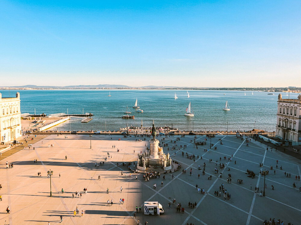 Panoramic view of Commerce Square in Lisbon. There are people on the square, a statue in the middle and the Tagus River.