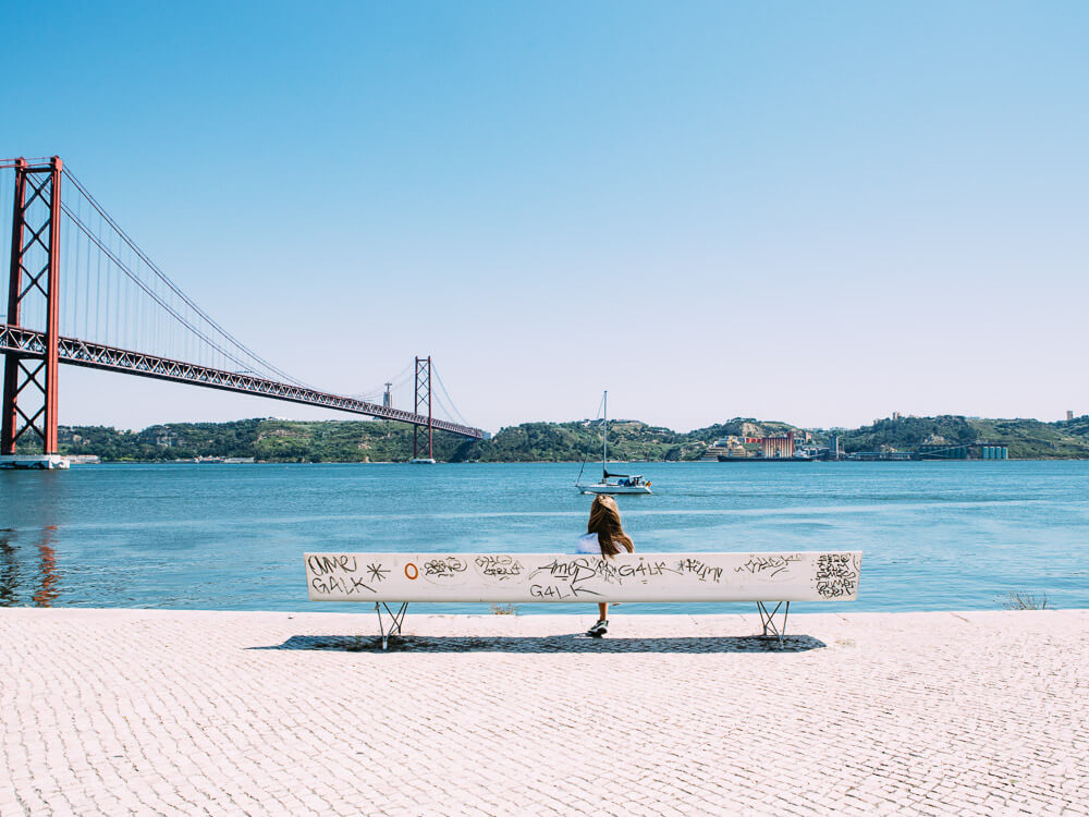 A woman enjoying the view of the Tagus River and bridge from a bench in Lisbon.