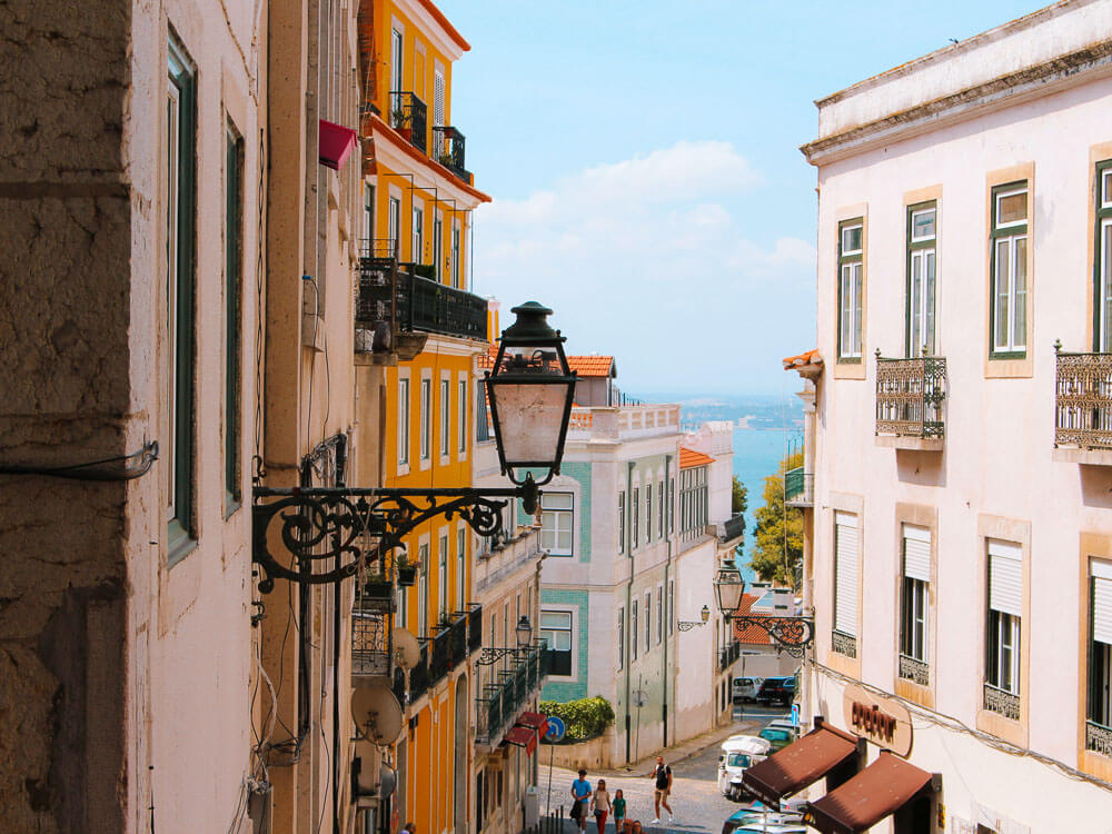 View of a street in Lisbon. There's a lamppost, an yellow building and people walking in the street. The Tagus River can be seen in the background.