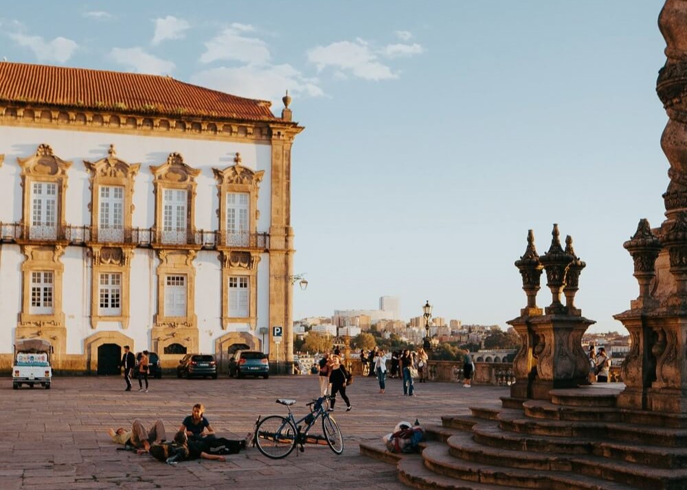 People sitting on the floor next to a bike in a square in Porto. There are stairs to the right and behind them there's a white and yellow historical building.