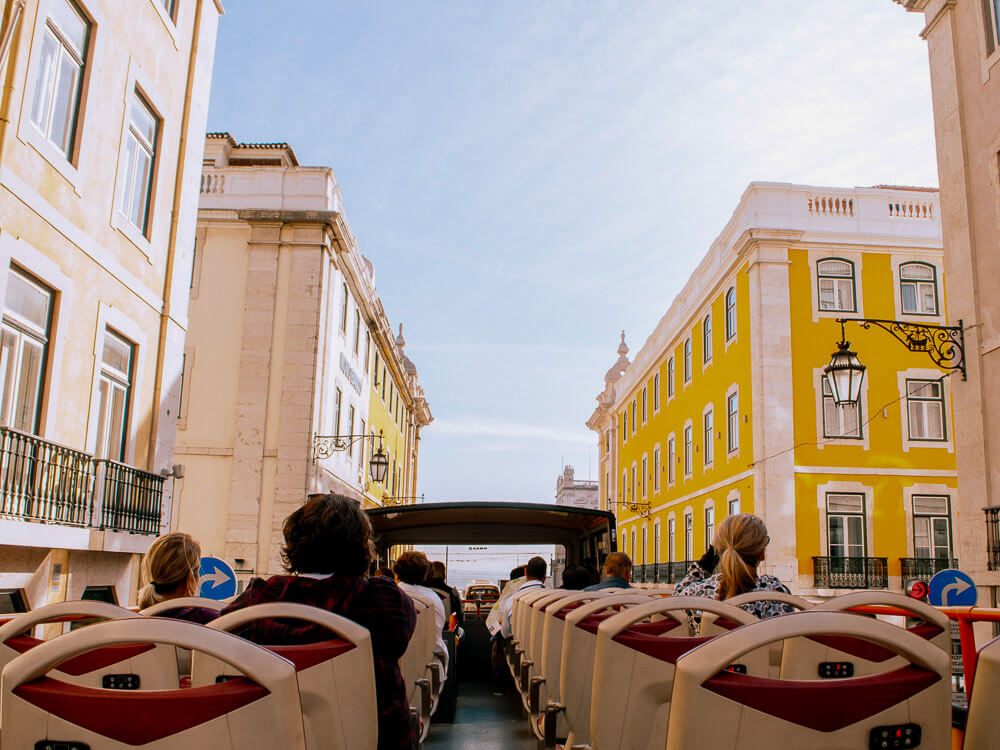 A group of people from the back. They are sitting in a tourist bus in Lisbon, looking at buildings.