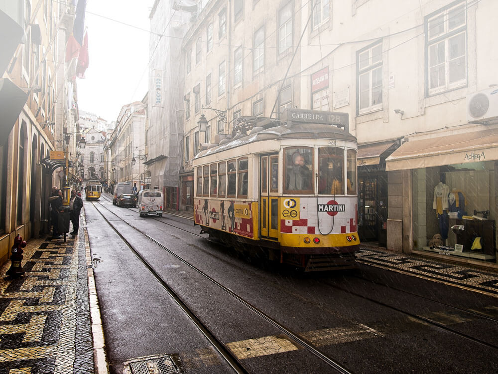 A street in Lisbon during the rain, with some fog. There's a traditional yellow and white tram in motion.