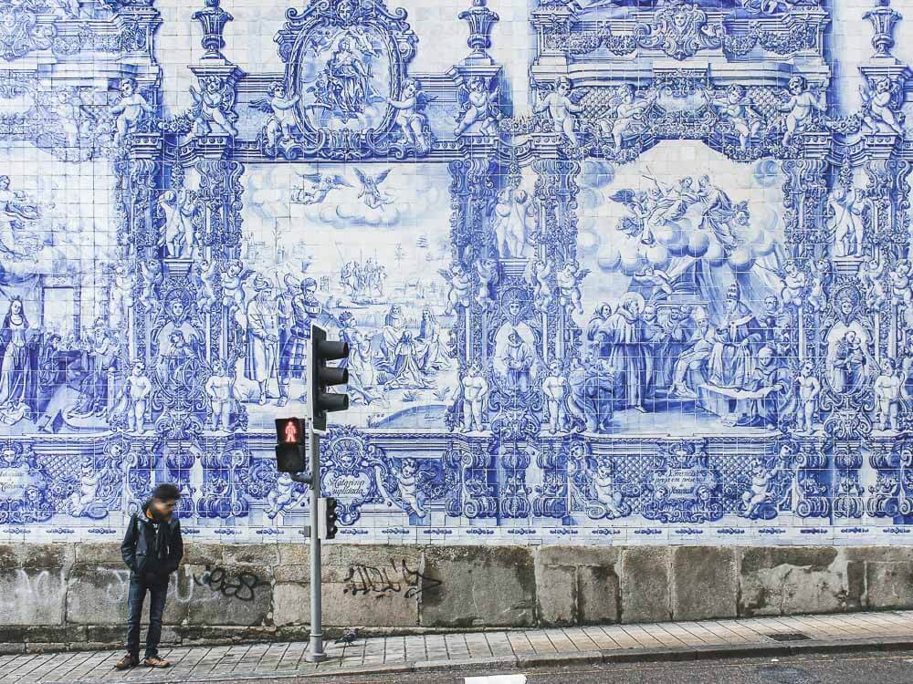 A man standing in front of a big mural of Portuguese tiles. Next to him there's a stop light.