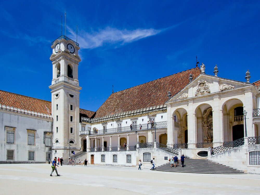 A tower with a large clock on top, standing tall beside a large white building in Coimbra, Portugal. 