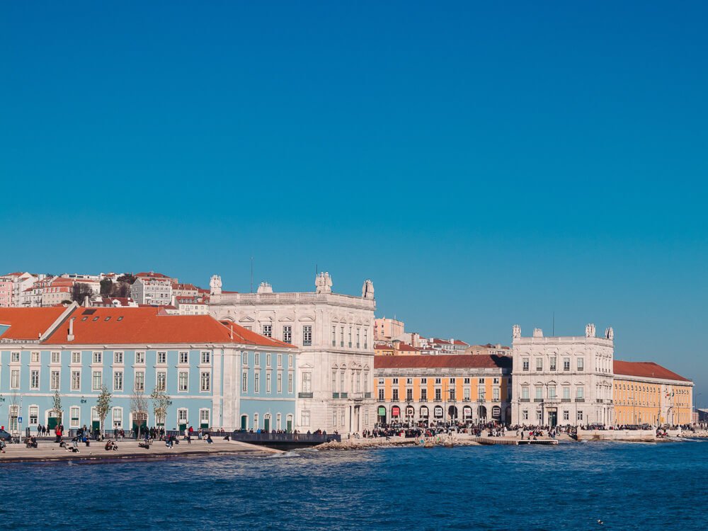 View of the Tagus riverside in Lisbon, with a blue construction with red roof to the left, a white one in the middle and a yellow one to the right. There are a lot of people near the margins of the river.