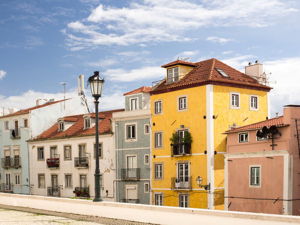 Colorful houses lining a street in Lisbon. From right to left, there’s a pink house, then a yellow, a blue, a white and another blue house. There’s a lamppost on a small wall. The sky is blue with some clouds.