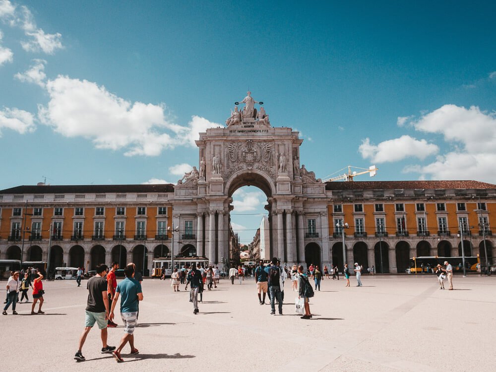 View of Commerce Square, in Lisbon. There’s a white concrete floor, where people are walking. You can see the Augusta Arch in the middle, with yellow constructions to each side of it.