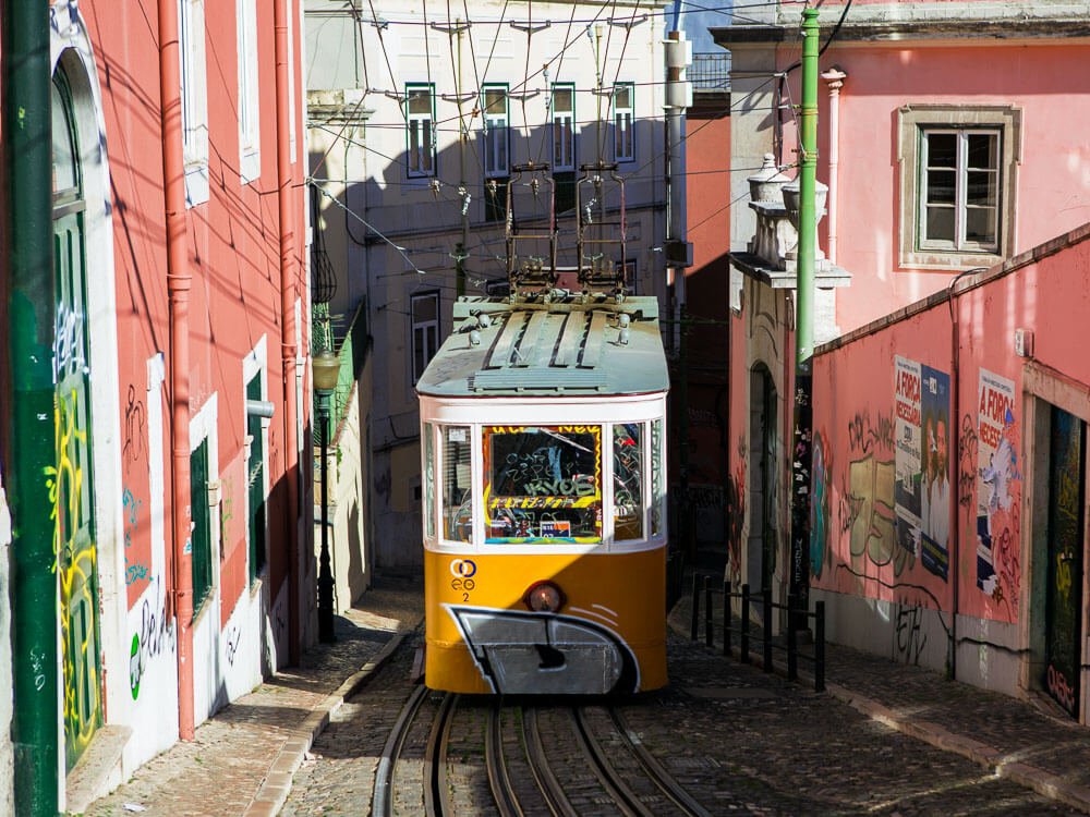 A yellow and white tram going up a steep hill in Lisbon during the day. On both sides, there are pink houses. 