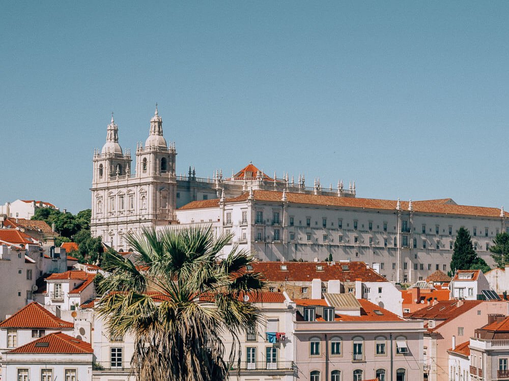 White buildings with red roofs in the Alfama neighborhood, in Lisbon. There’s a big white construction in the middle, with small towers surrounding it. The sky is blue.