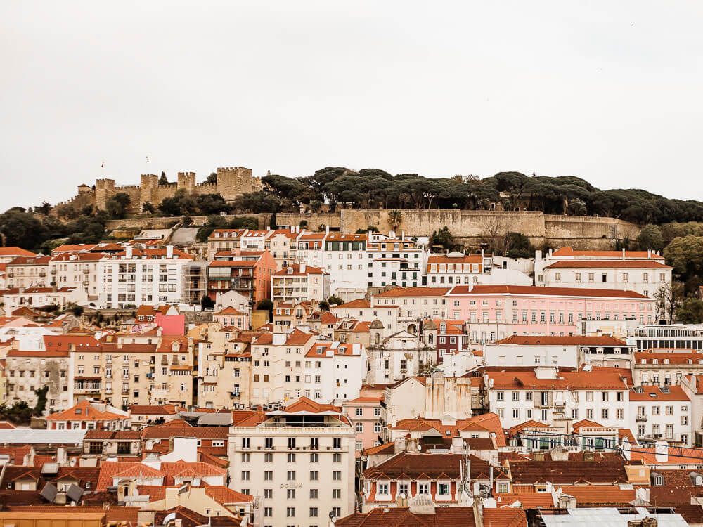 View of Alfama with St. George’s Castle above. Beneath it, there are numerous concrete houses with red roofs. The sky is grey.