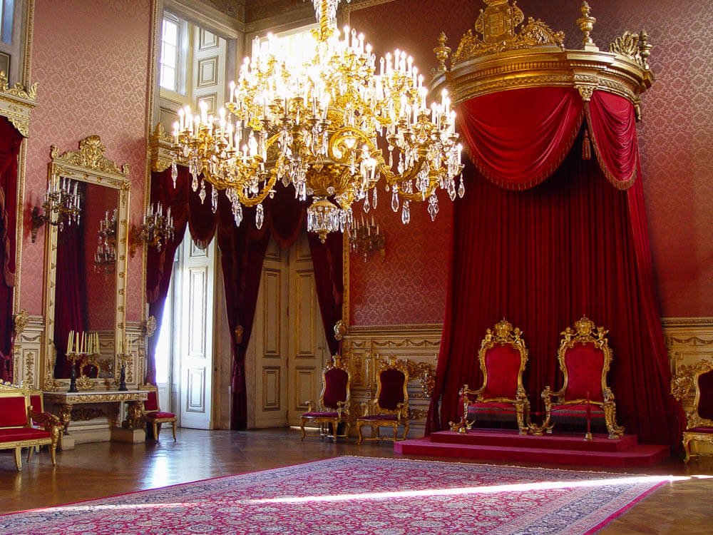 The Throne Room in the Ajuda National Palace. The walls have red wallpaper, and at the bottom, there are golden details with a white background. There is an ornate red carpet on the floor and a sumptuous chandelier in the middle. Two thrones are on a small platform, with a red curtain behind them.