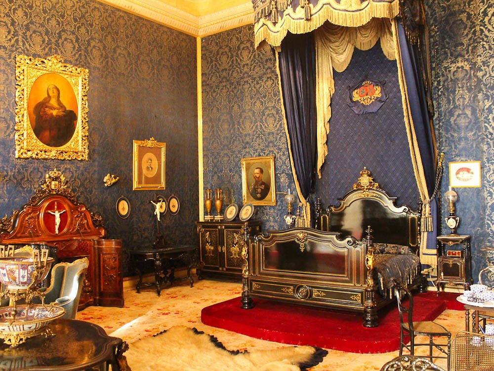 A bedroom with ornate blue and gold wallpaper. A black and gold bed with a blue and gold canopy on a red platform. There are paintings hanging on the walls, a fur rug, and various period furniture.