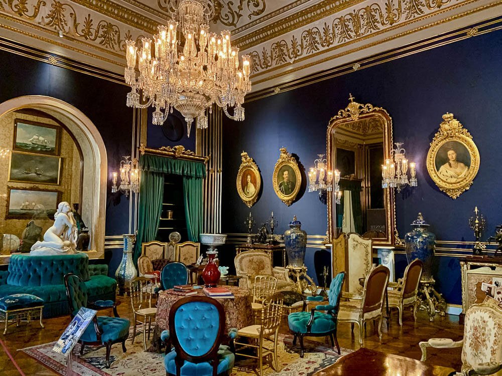 A sumptuous room in the Ajuda National Palace. The walls are blue, and the ceiling is white with golden ornaments. There is a chandelier in the middle. One of the walls has paintings in golden round frames, and a mirror with a lamp on each side. There is a statue on top of a round sofa on the left side. There are many different chairs scattered around the room.