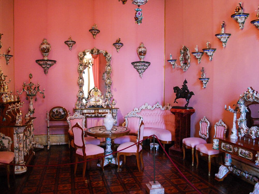 A room with pink walls and various porcelain hanging. In the center, there is a table with four chairs with pink upholstery. To the right, a corner sofa, a horse statue on a pedestal, and an ornate dressing table.