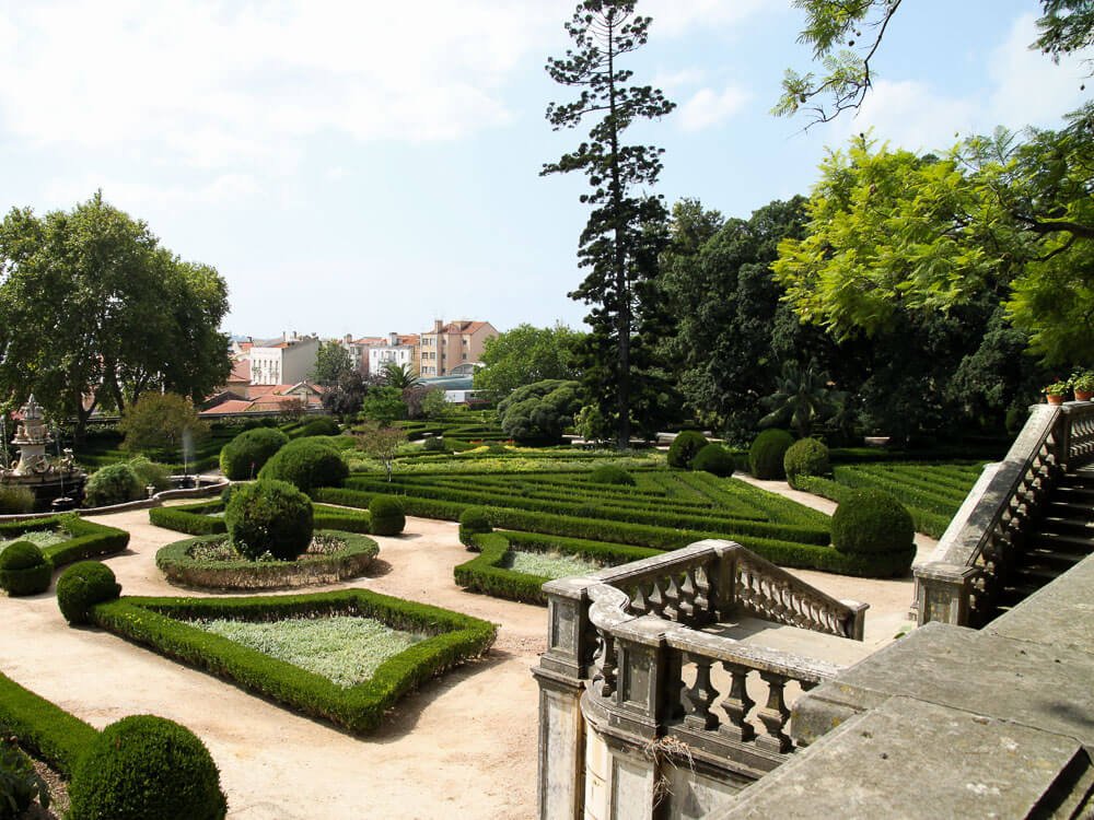 Palace gardens, with trimmed bushes and various trees surrounding. There are dirt paths, and on the right, a staircase. In the background, the city of Lisbon.