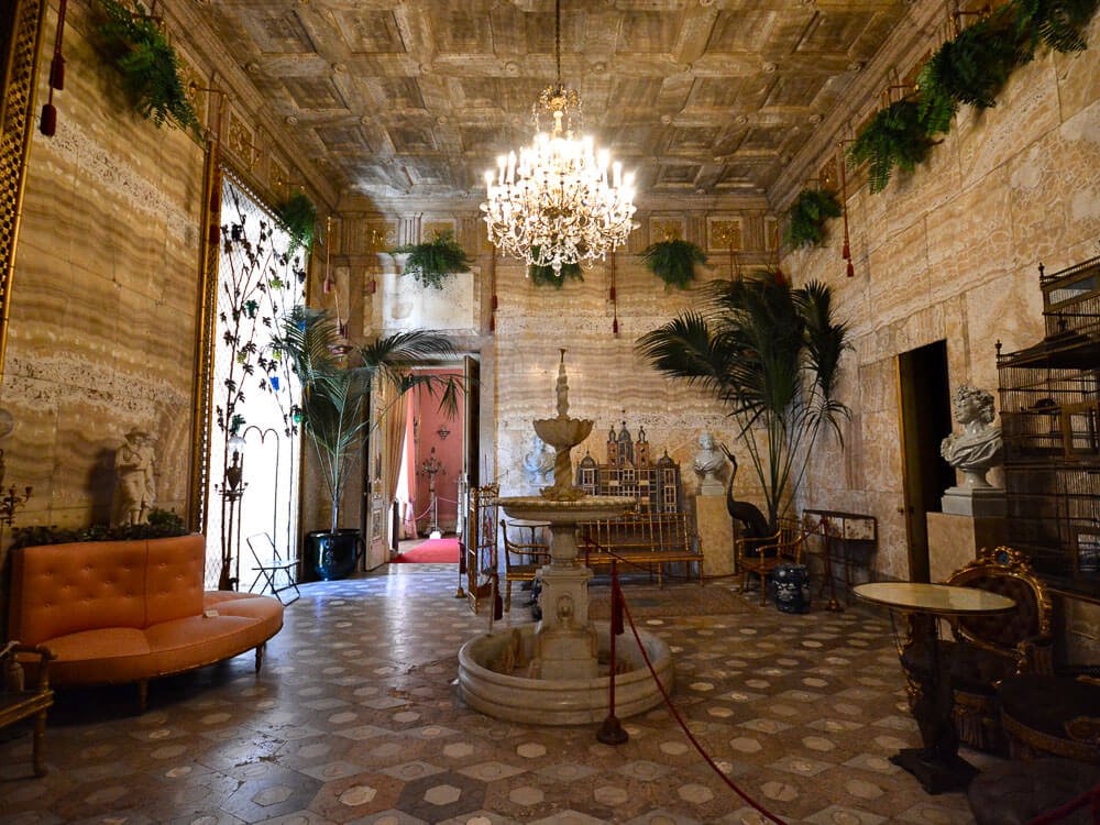 A room with marble walls in the Ajuda National Palace. There is a fountain in the middle and a chandelier above it. On the right side, there are cages, a bust, and a table. On the left side, a sofa. There are plants in the background and near the ceiling.