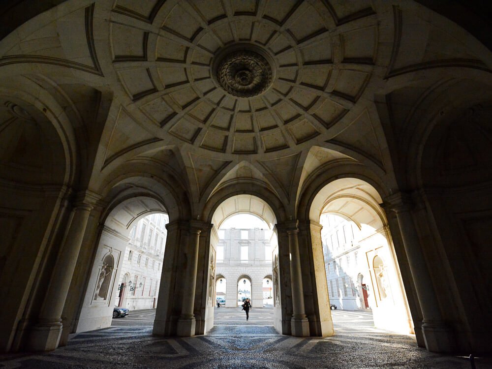 Part of the architecture of the Ajuda National Palace. A high-ceilinged vaulted construction, with three arches leading to an illuminated courtyard. In the middle, a person in black.