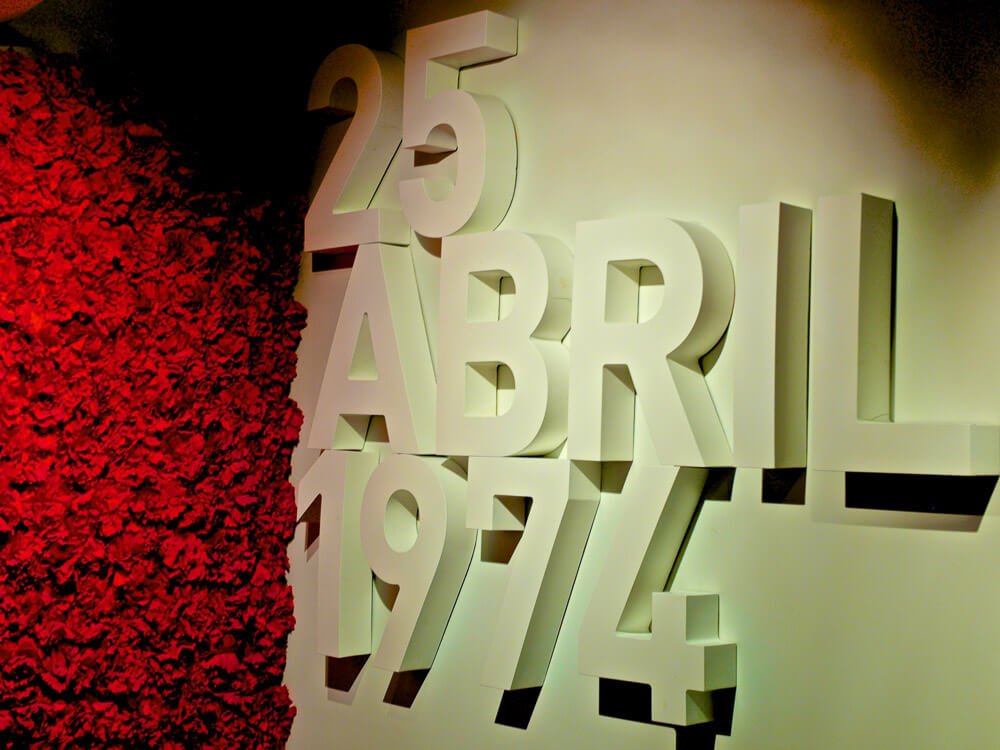 A white wall in the Aljube Museum, with "25 de Abril 1974" written in white raised letters. On the left side, a wall with red carnations, alluding to the Carnation Revolution.