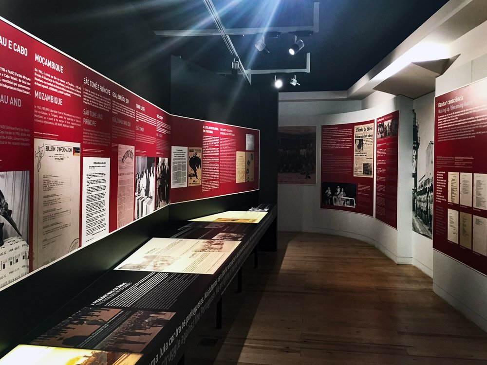 A wooden-floored corridor in the Aljube Museum. On the left side, a black wall with a red panel featuring photos and texts in white letters. On the right side, a white wall also with a red panel containing written information.