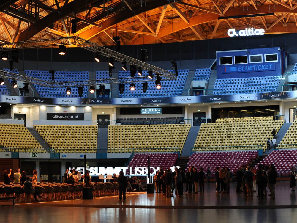 Interior of the Altice Arena. At the top, there are blue chairs. A little further down, there are yellow chairs, and even further down, there are red chairs. There is a group of people standing in the middle, and chairs arranged on the floor to the left.