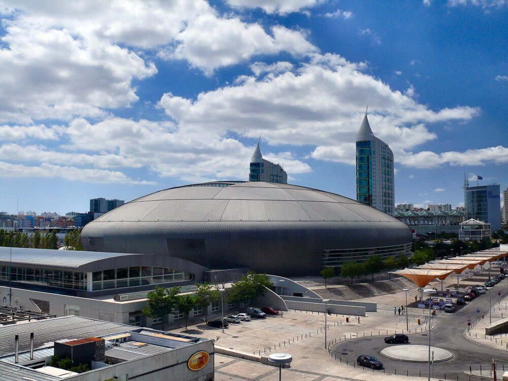 View of the exterior of the Altice Arena against a blue sky with some clouds. Behind, there are some buildings, and on the right side, a parking lot with cars.