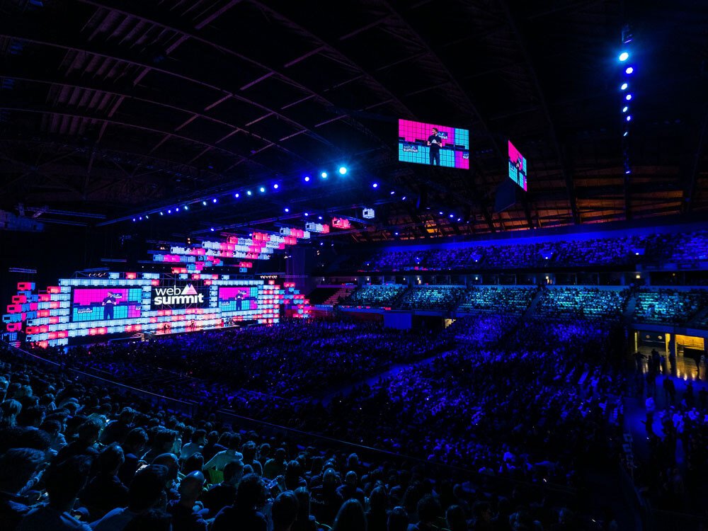 Interior of the Altice Arena, with blue lighting and many people seated. On the main screen, it says "Web Summit" and there are two smaller screens near the ceiling.