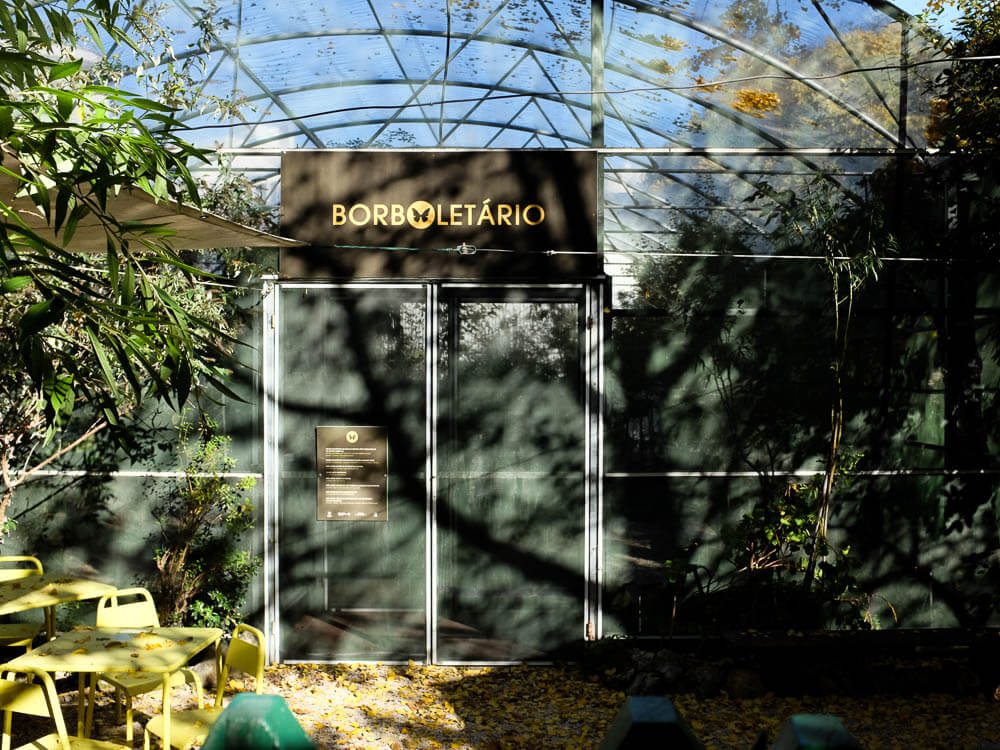 An iron structure surrounded by mesh at the Lisbon Botanical Garden, with a sign above double doors saying "Butterfly House." On the left side, a yellow table with chairs and a parasol. On the right side, shadows cast by trees.