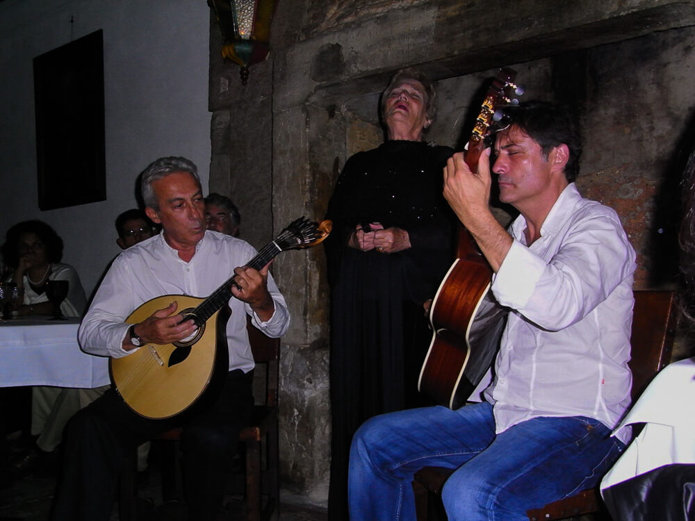 Two men in white shirts playing instruments and a woman with short hair dressed in black singing. Behind them, a gray stone wall. On the left side, a table with people watching the performance.