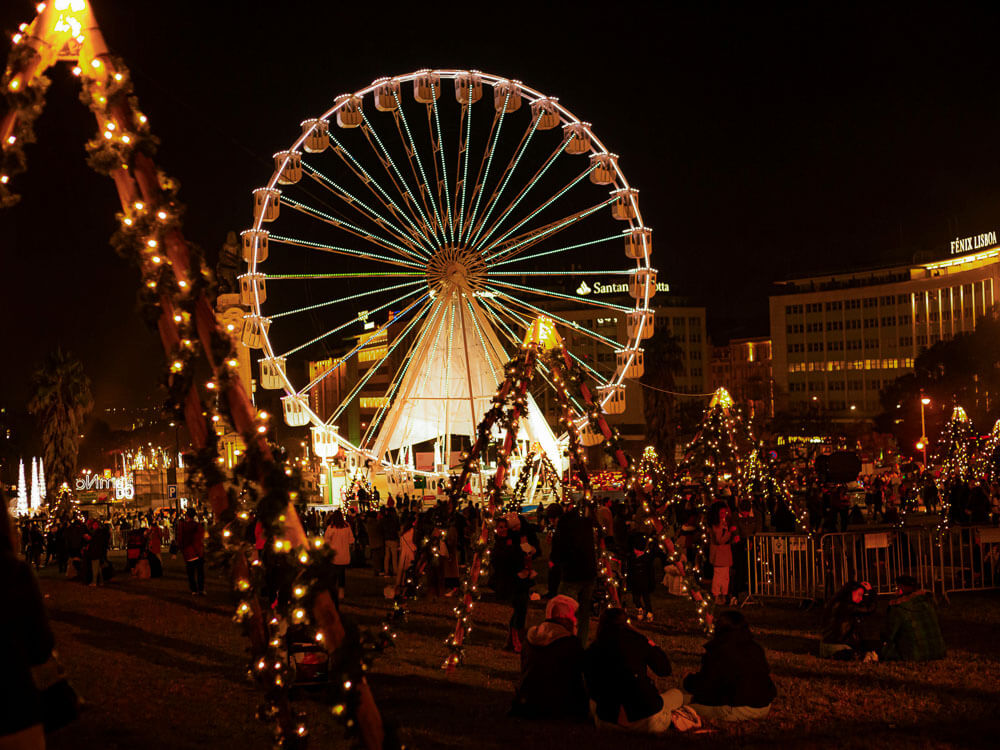 Christmas decoration in Lisbon in December. There are triangular structures with Christmas lights, people sitting on the ground, and scattered around an illuminated Ferris wheel.