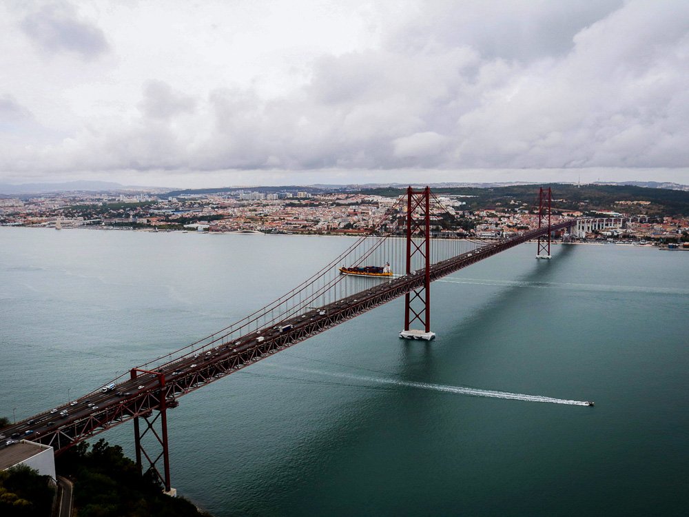 A red bridge in Lisbon in December, across the Tagus River. The sky is cloudy and you can see the city in the background.