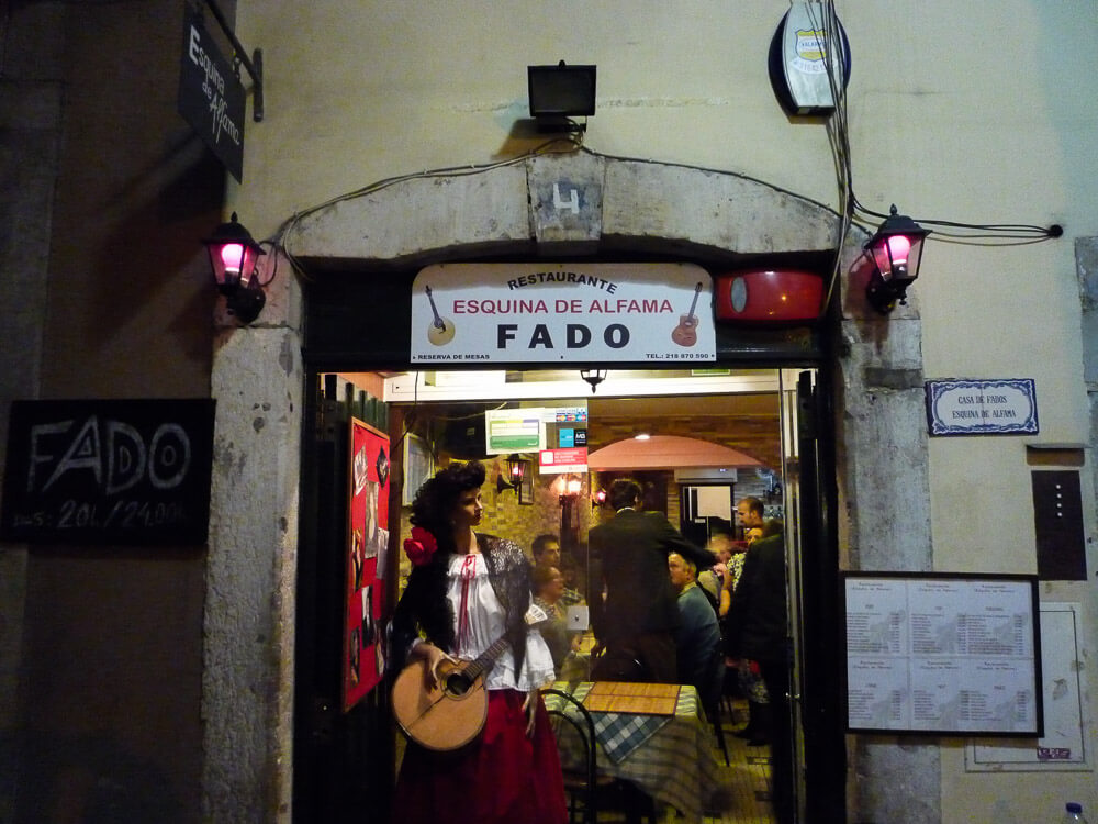 Entrance of a restaurant in Alfama, with an arch and a red light fixture on each side. A sign above says "Restaurante Esquina de Alfama Fado." On the left side, a sign that says "fado." On the right side, the menu. Inside, there is a mannequin holding a musical instrument. Behind it, several people gathered and talking.