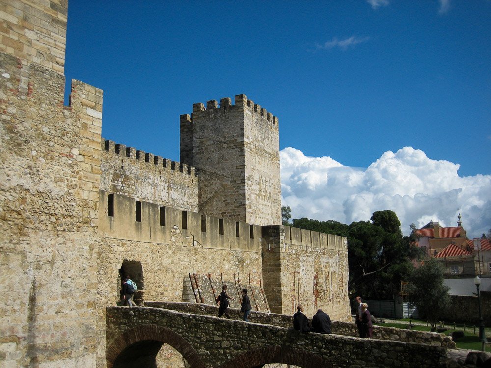 Entrance of São Jorge Castle. An old stone bridge and two stone towers, with people entering.